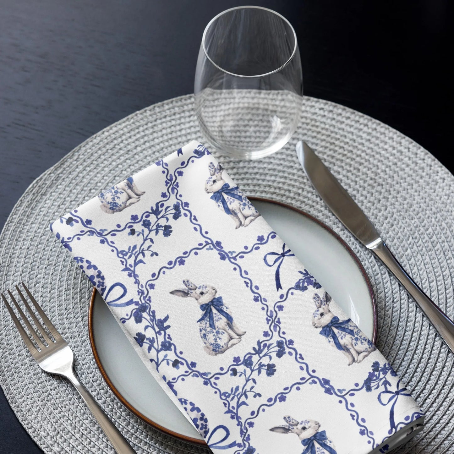 White napkin with a blue Chinoiserie rabbit and floral design displayed on a set table with a plate, silverware, and glass.