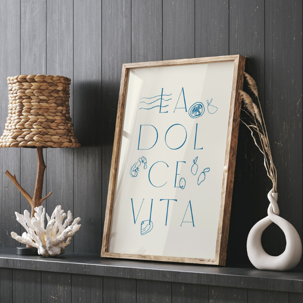 La Dolce Vita print leaning on a dark wood shelf with natural coastal decor, seashell motif and blue summer illustrations in frame