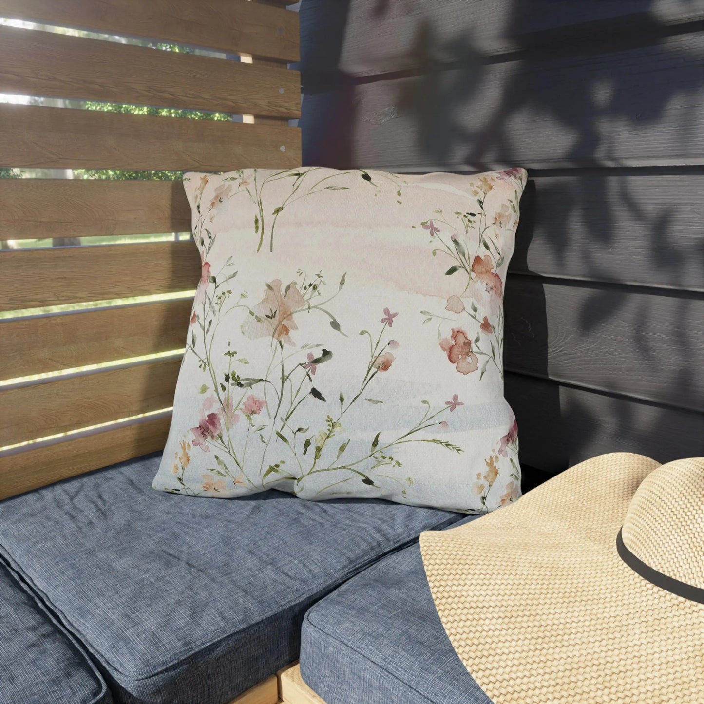 Outdoor pillow with soft watercolor pink wildflowers and green stems, styled on a patio bench in dappled sunlight
