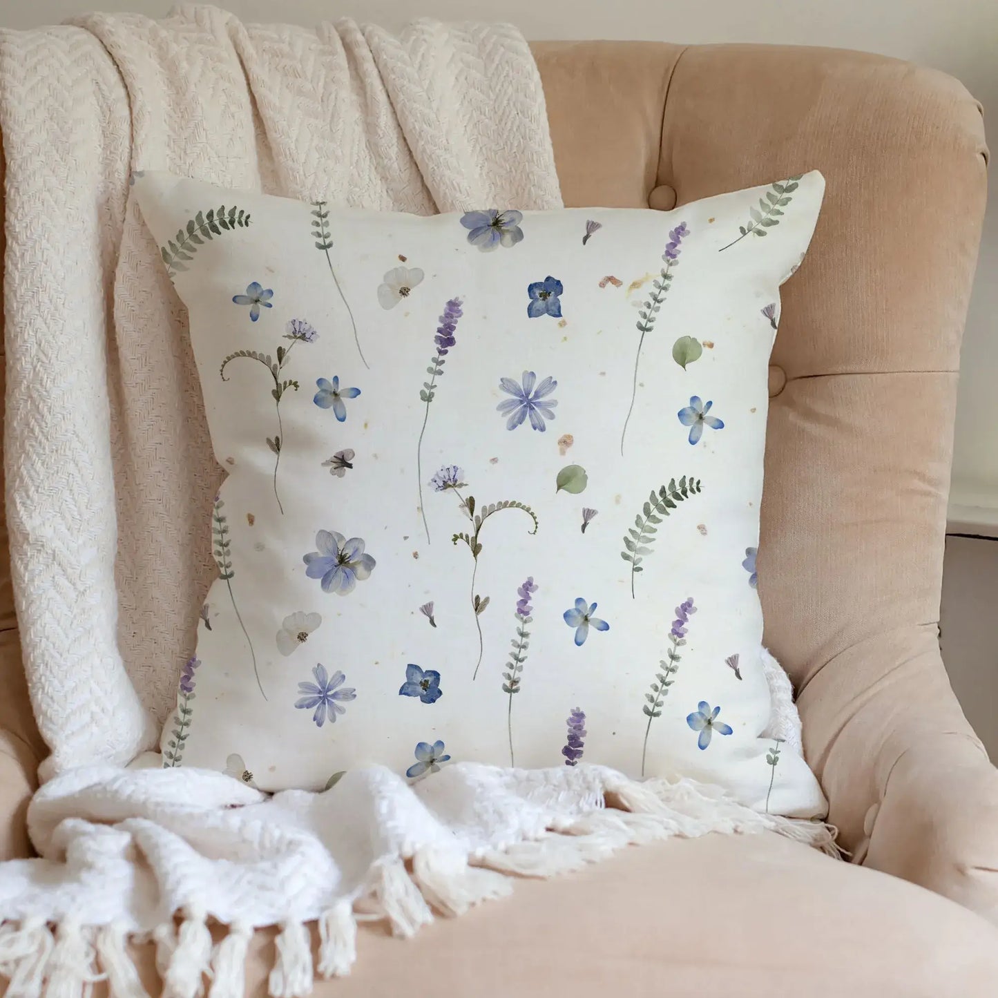 Cream throw pillow with blue and purple pressed flower design on a textured armchair. Nature-inspired living space.