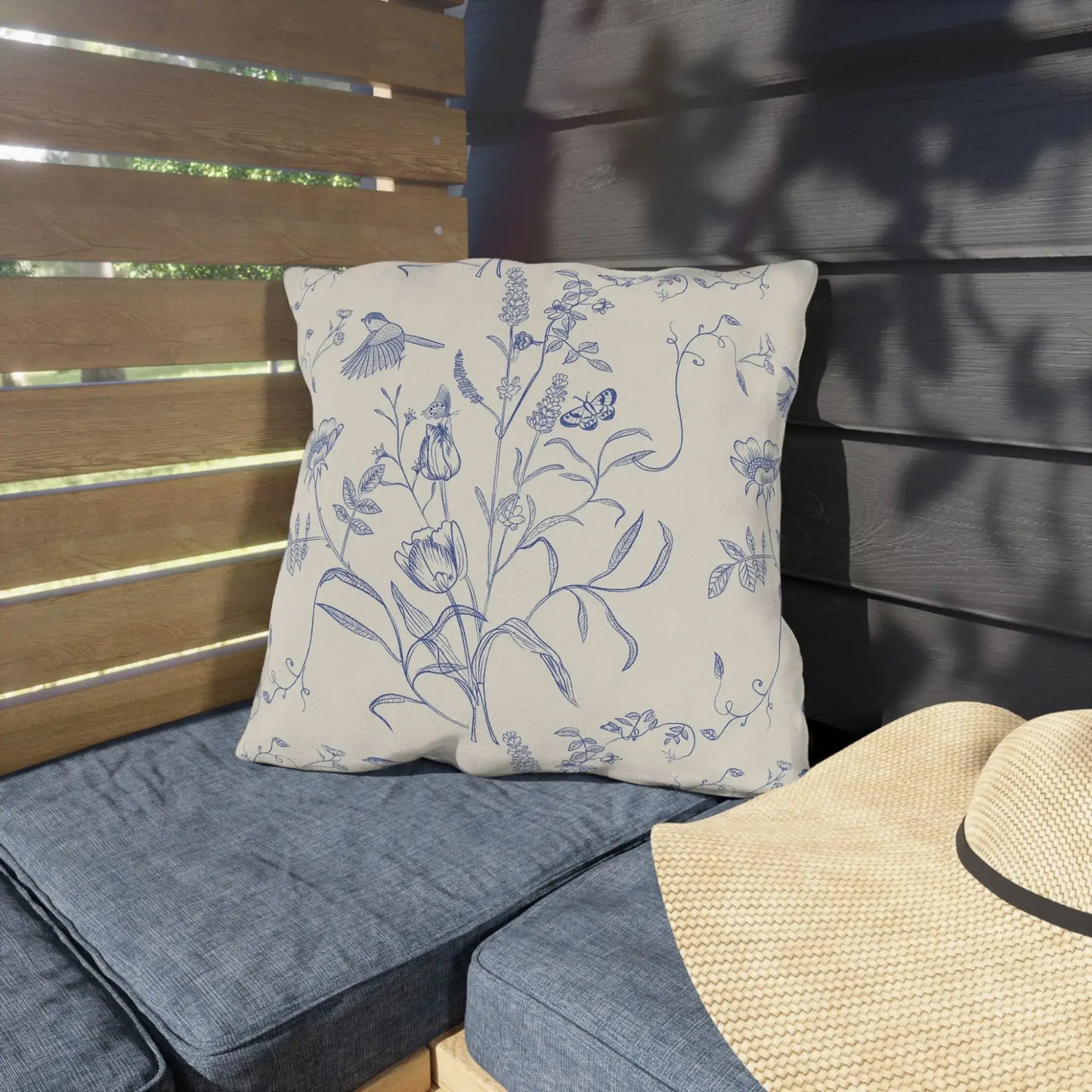 Blue and white botanical outdoor pillow with birds and butterflies, displayed on a patio bench with denim blue cushions and a woven sun hat — garden-inspired design.







