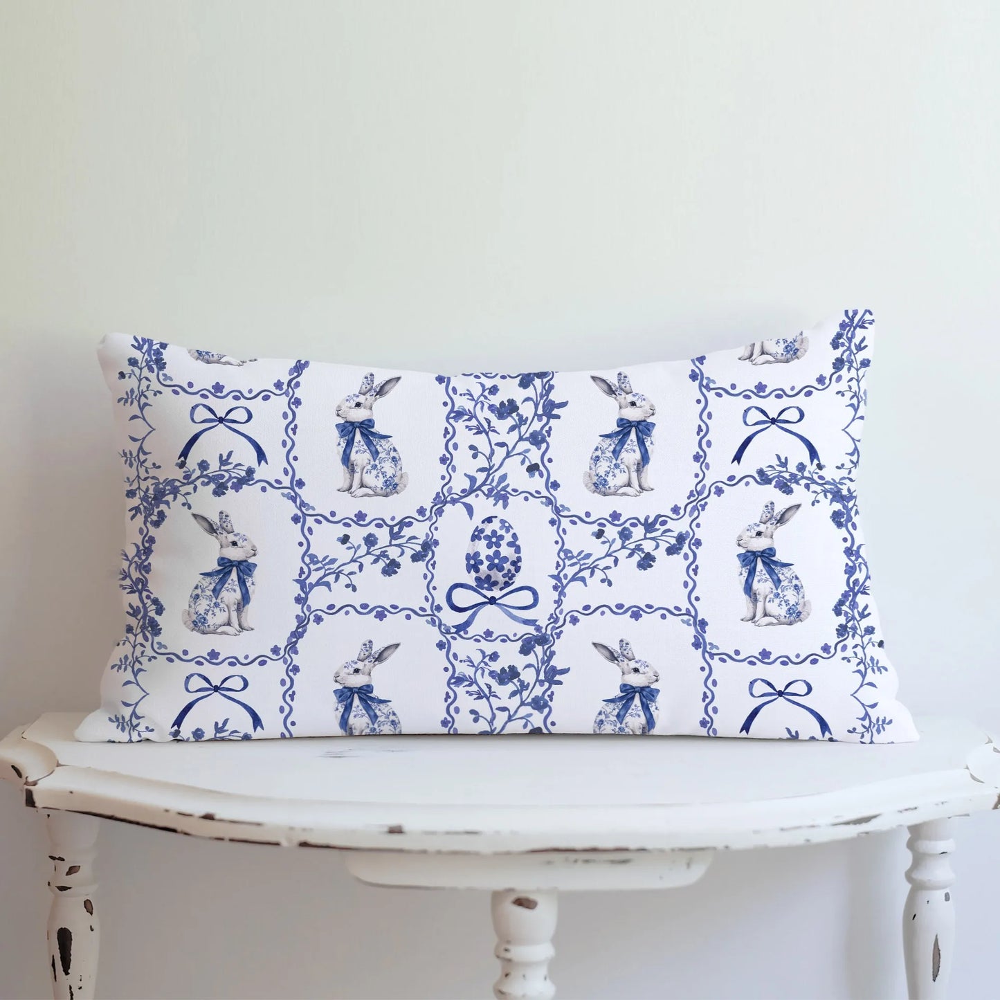 Lumbar pillow with a blue watercolor Chinoiserie tile pattern featuring rabbits with bows and decorated Easter eggs, displayed on a white table.