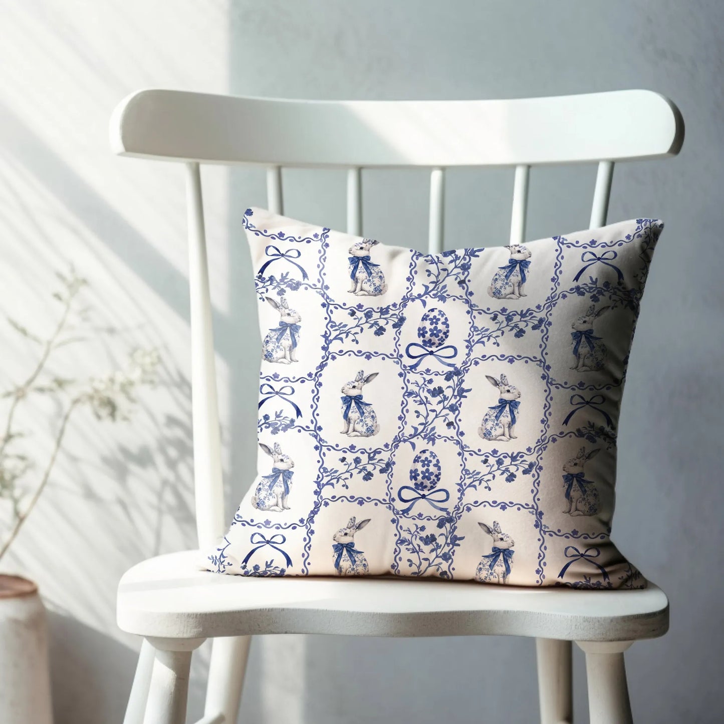 White pillow with a blue watercolor Chinoiserie tile design of rabbits with bows and Easter eggs, displayed on a white chair.