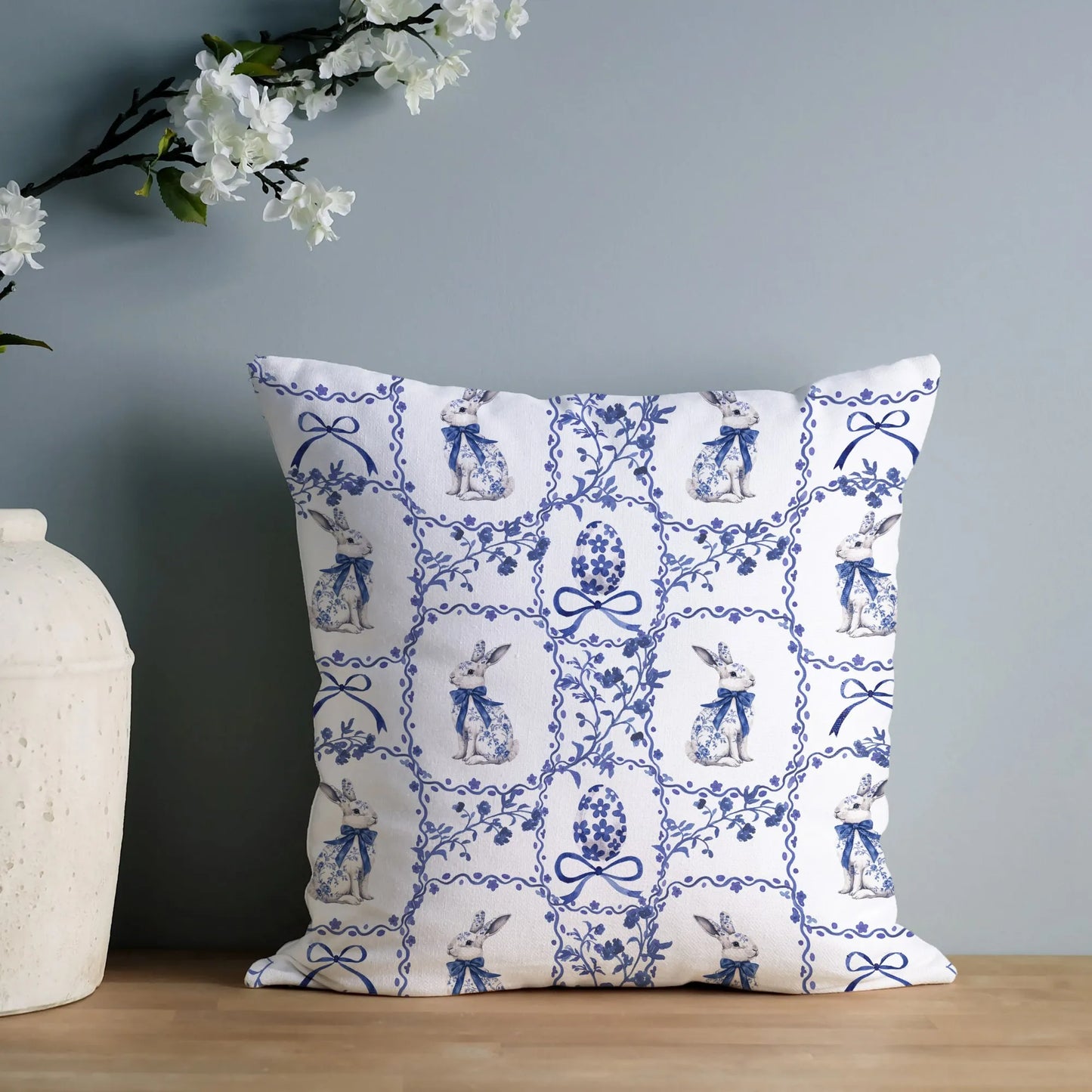 White pillow featuring a blue watercolor Chinoiserie tile pattern with rabbits and Easter eggs, styled with white blossoms.