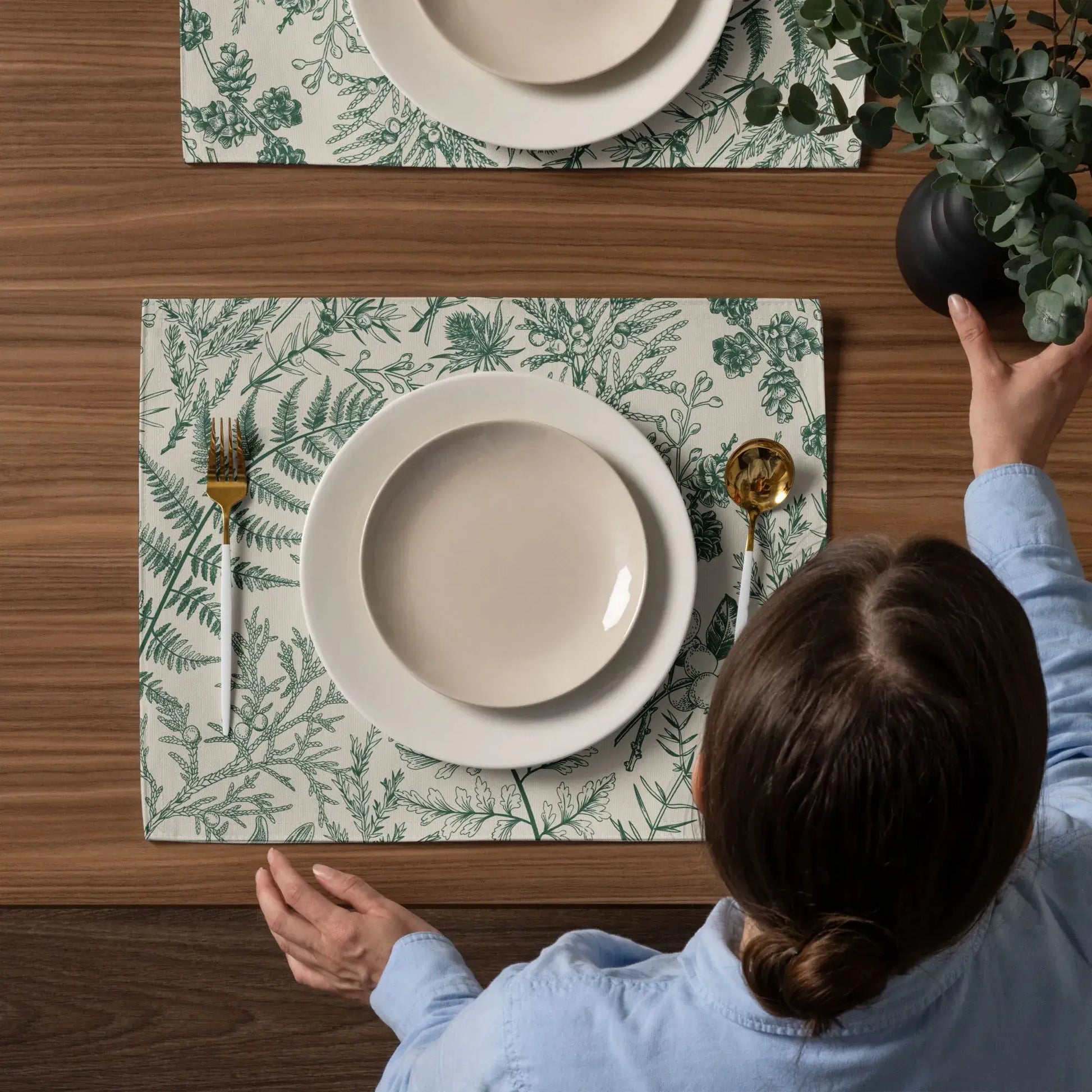 Botanical Christmas placemats set on a wooden table with plates and utensils, showcasing winter greenery decor.