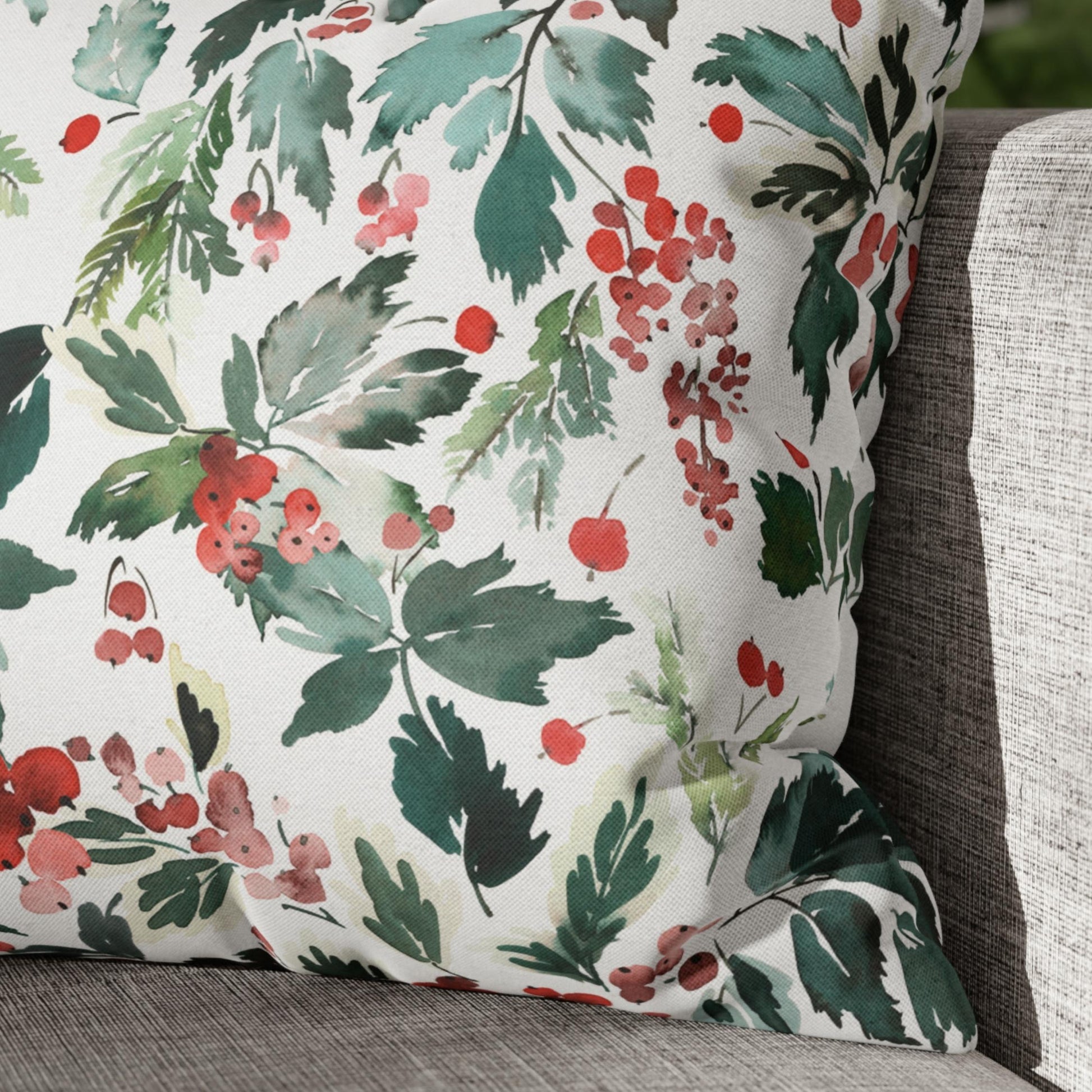 Close-up of watercolor Christmas pillow with holly berries and leaves on gray couch – festive botanical decor