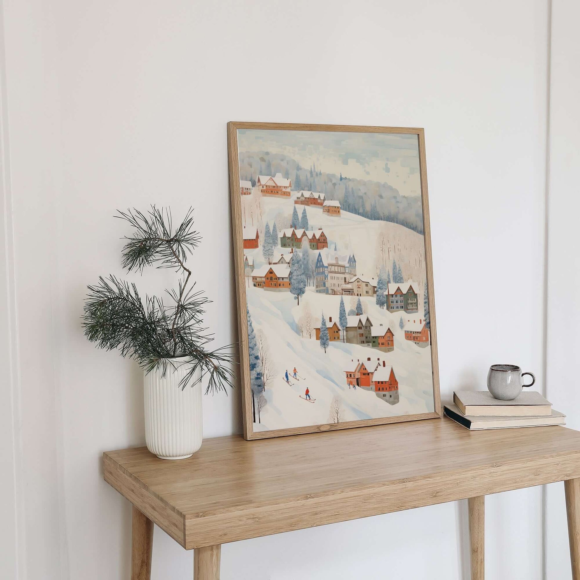 Cute Winter Village Art print displayed on a wooden table with a plant and books, showcasing a snowy village scene.