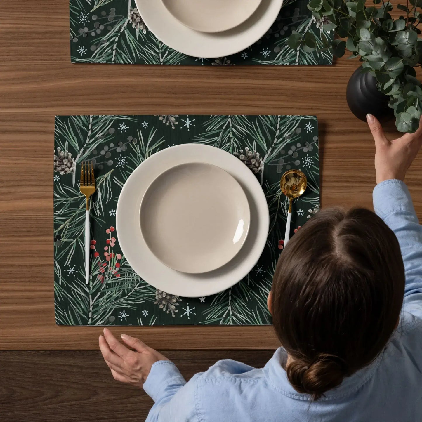 Person setting a table with Green Pine and Berry Winter Botanical Placemats and elegant tableware.
