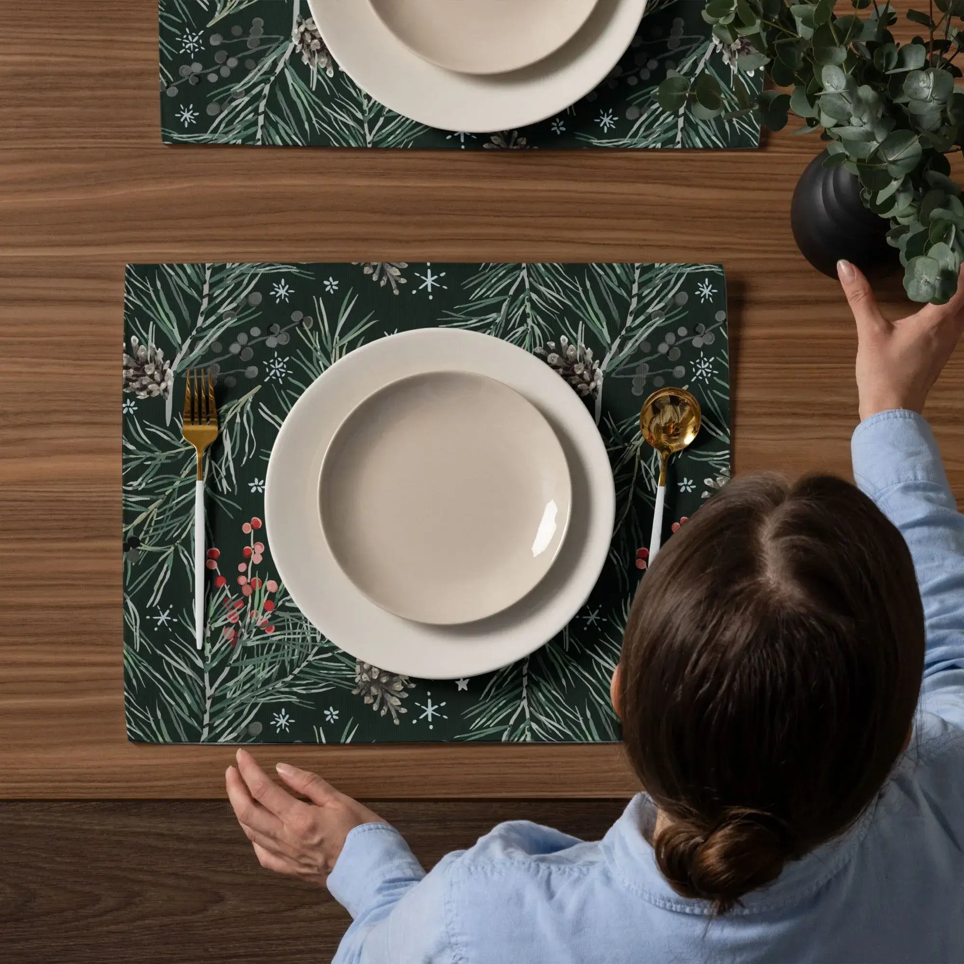 Person setting a table with Green Pine and Berry Winter Botanical Placemats and elegant tableware.