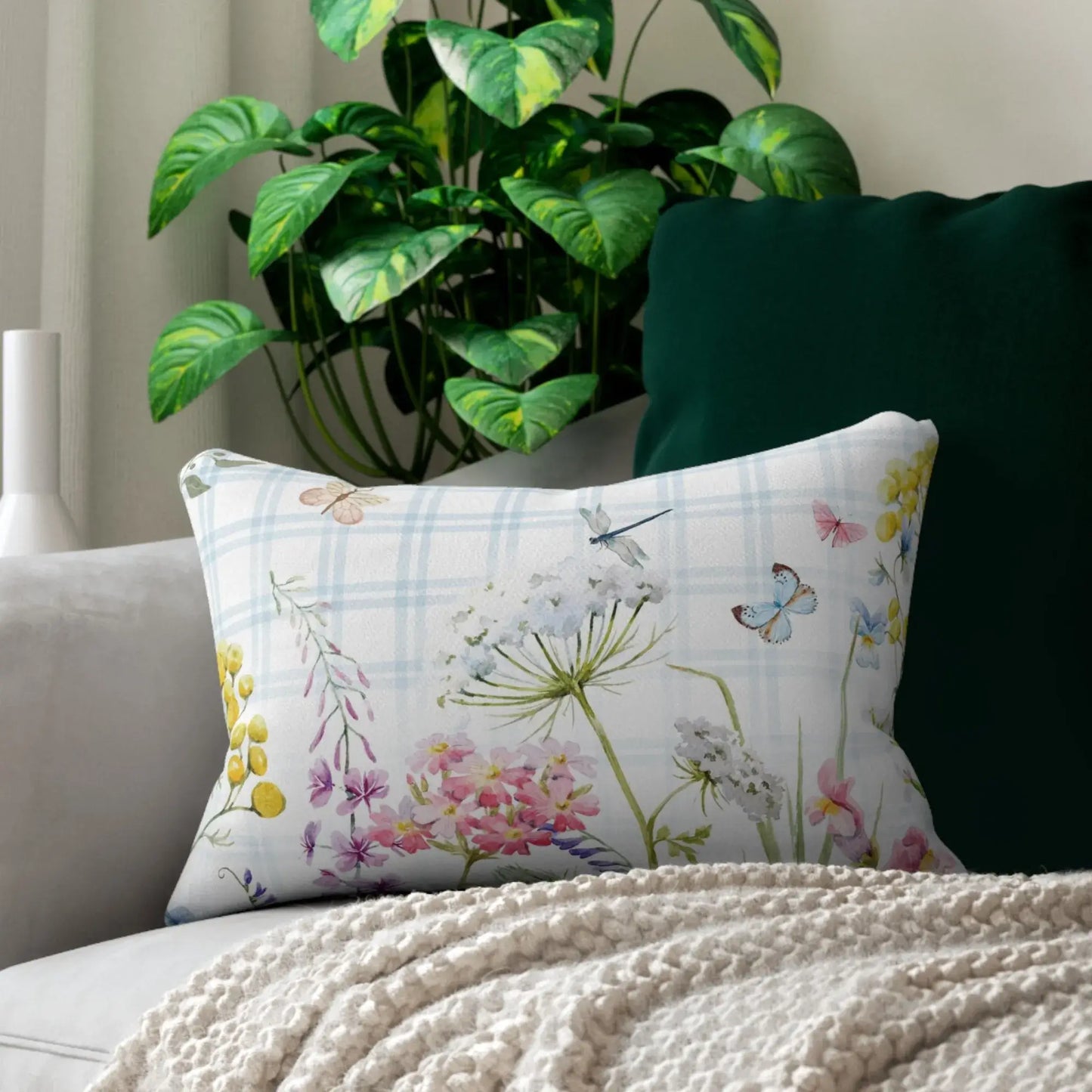 Watercolor lumbar pillow featuring delicate wildflowers, butterflies, and dragonflies on soft blue plaid — styled on a cream couch with cozy knit blanket and greenery.