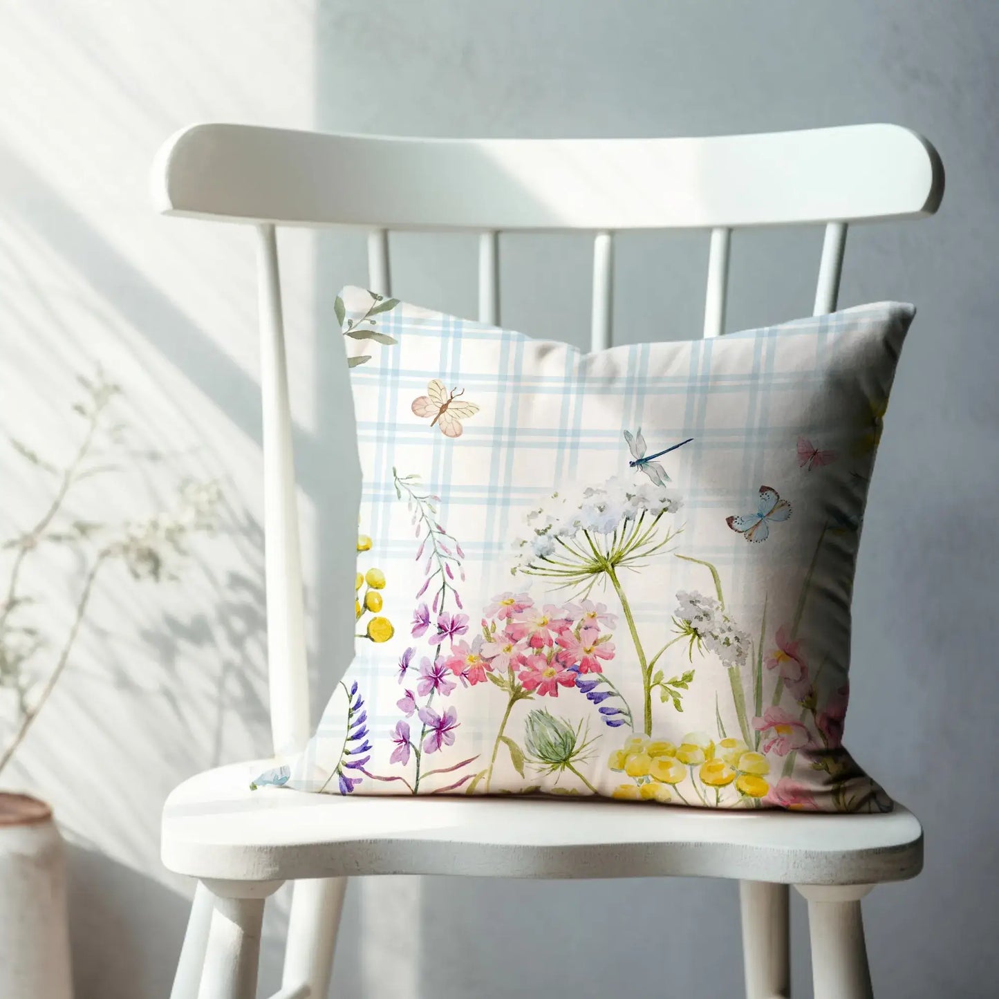 Watercolor floral throw pillow with pastel wildflowers and blue plaid background on white wooden chair — nature-inspired home decor accent.
