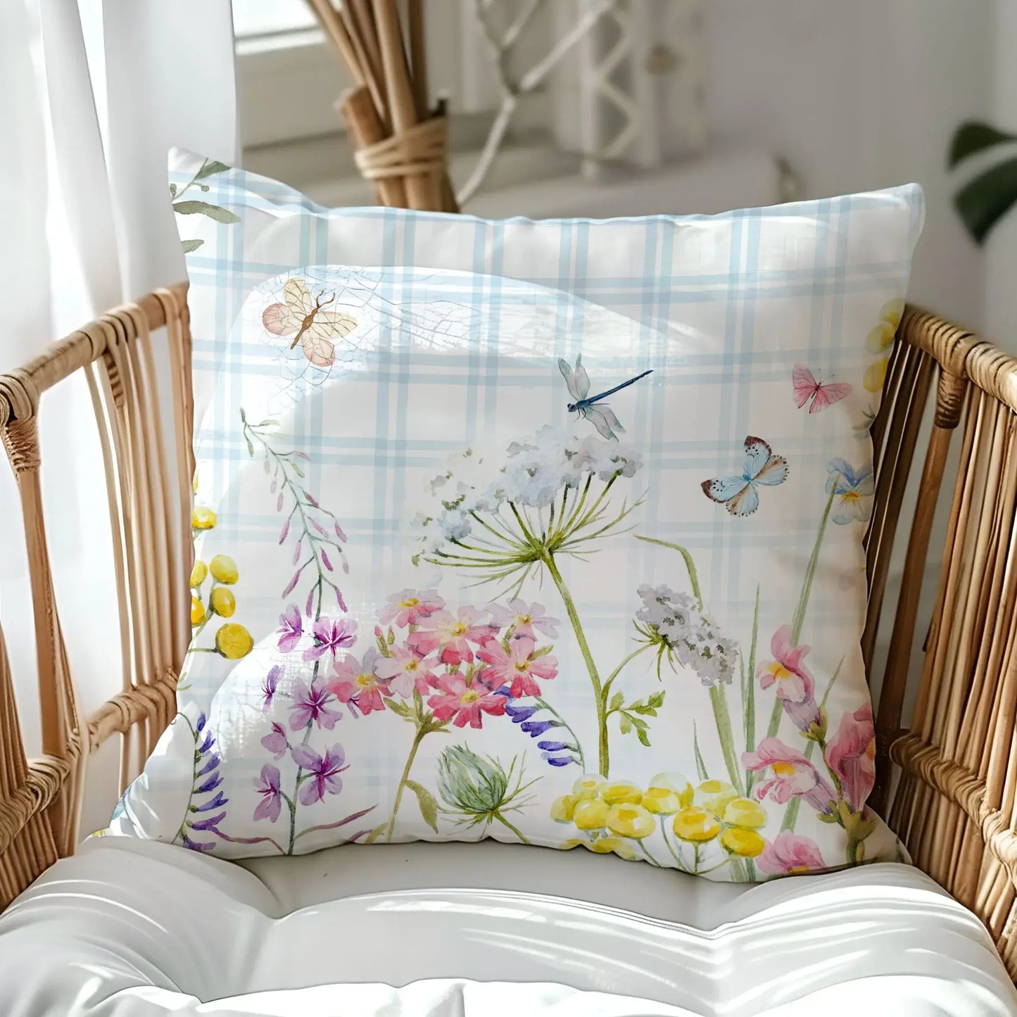 Watercolor floral outdoor pillow on rattan chair with pastel blue plaid background, featuring butterflies and wildflowers — nature-inspired spring decor.