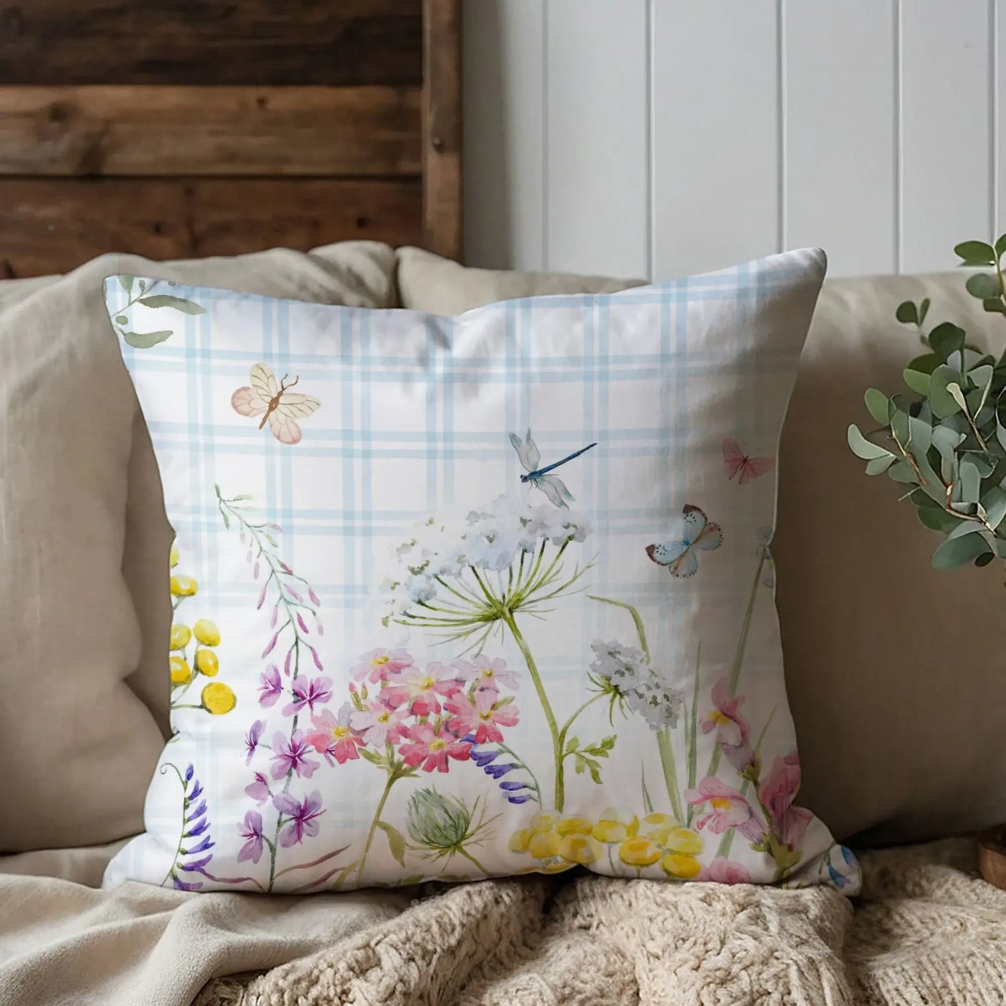 Watercolor floral pillow with pastel wildflowers and blue plaid background on neutral couch — cozy nature-inspired home decor accent.