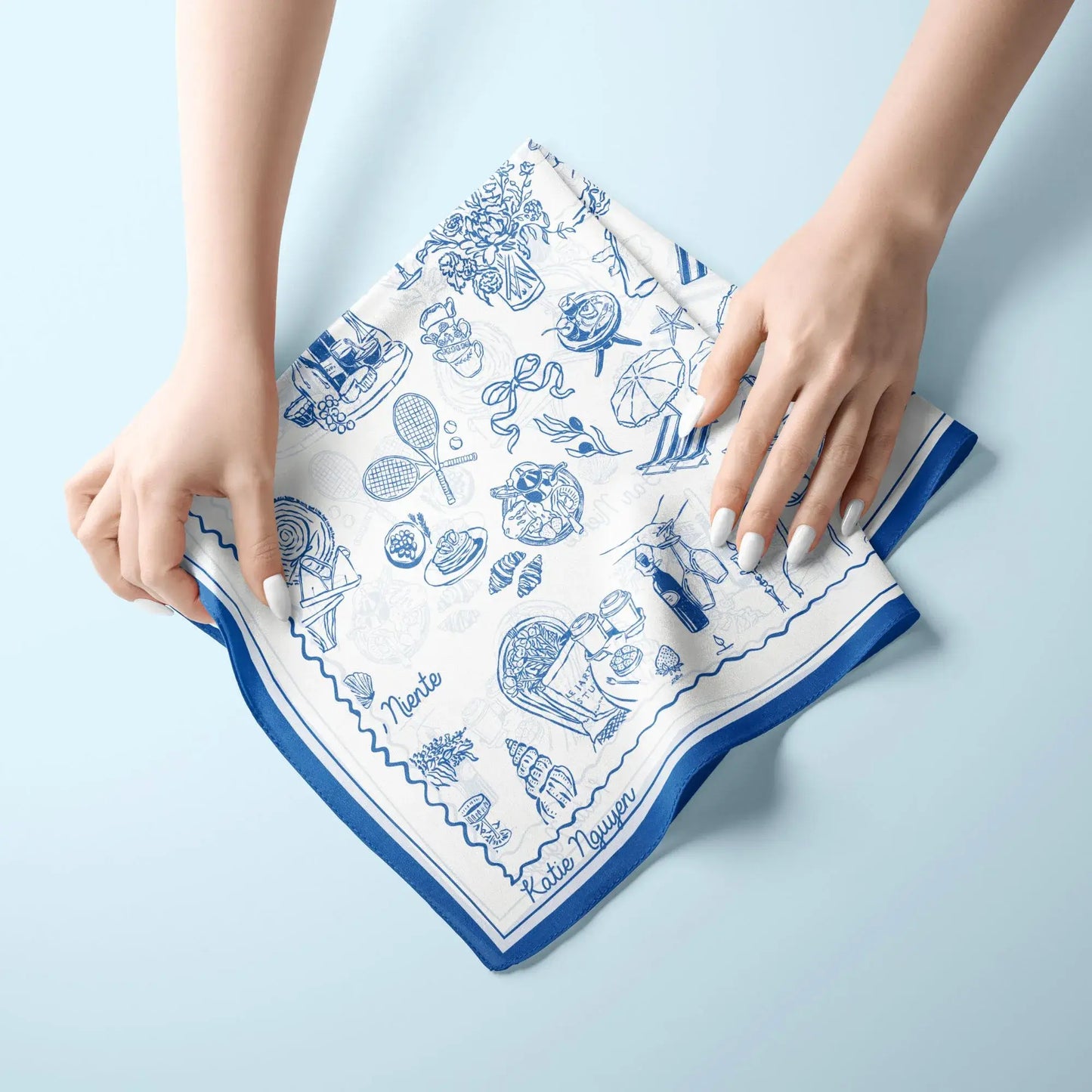 The Dolce Moment Silk Scarf in blue – hands folding silk square with Mediterranean summer illustrations.