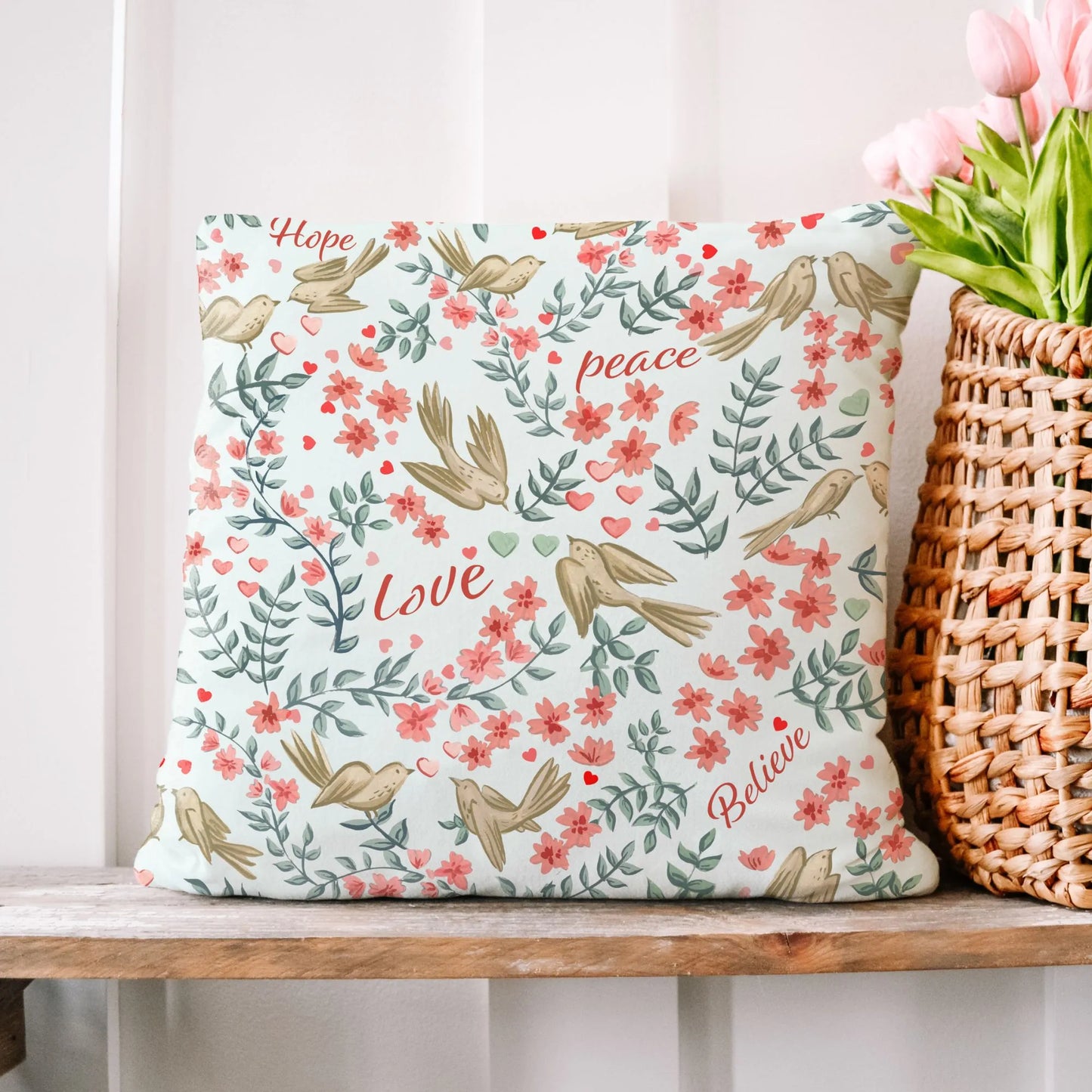 Square Love Birds Valentine Pillow Cover with "Hope Peace Love Believe" text, bird and floral design, resting on a wooden shelf beside a woven basket of pink tulips.