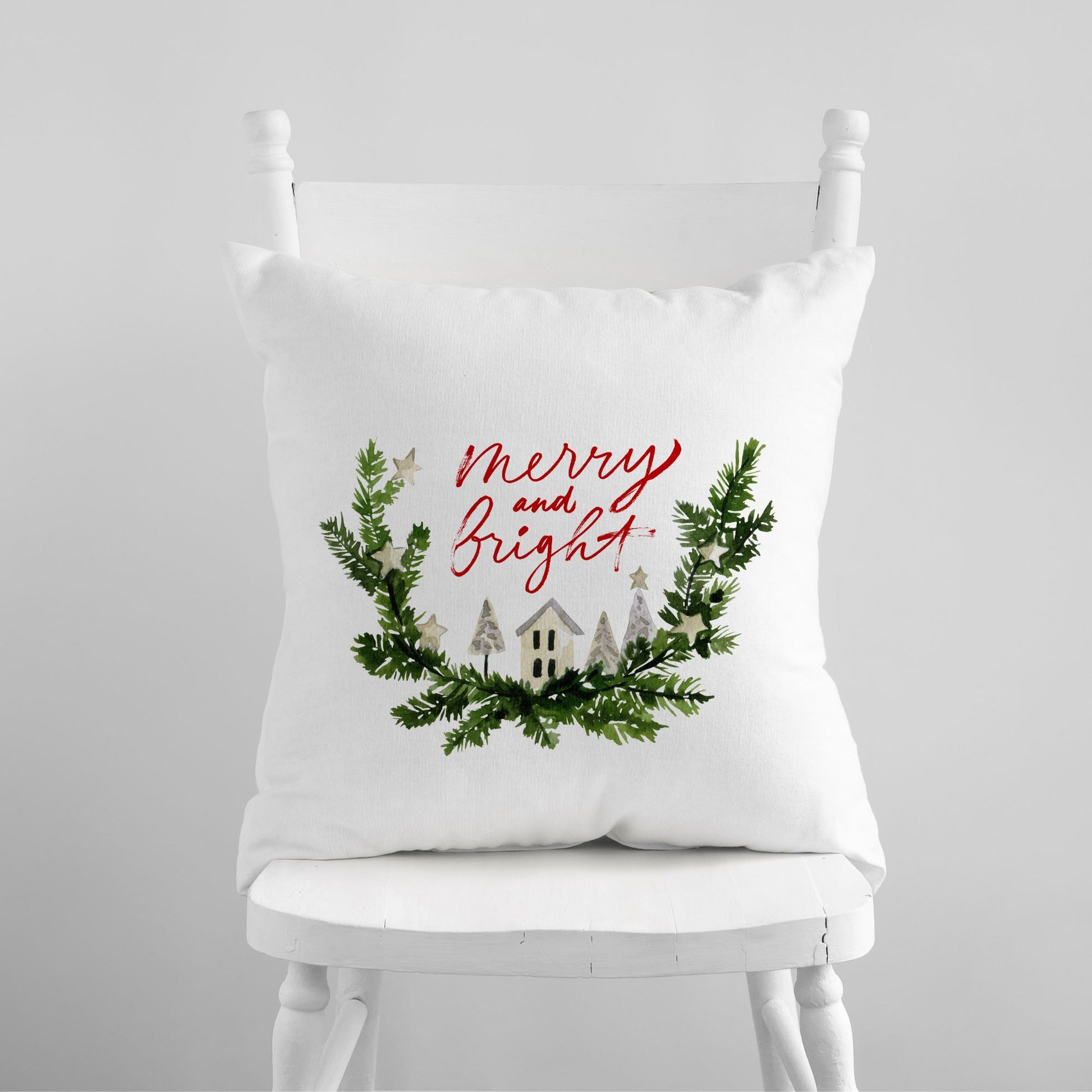 White pillow with "Merry and Bright" red script and watercolor winter village wreath – festive holiday decor