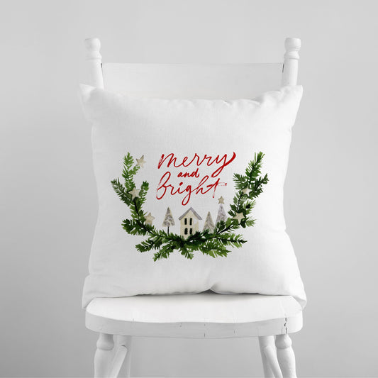 White pillow with "Merry and Bright" red script and watercolor winter village wreath – festive holiday decor