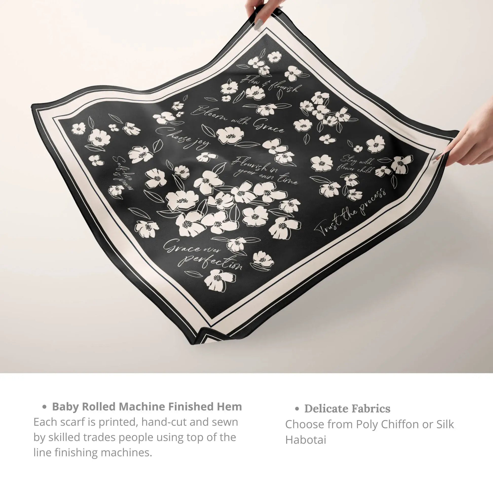 Hands holding a black silk scarf with white wildflower and affirmation design, showing the delicate hem and fabric.