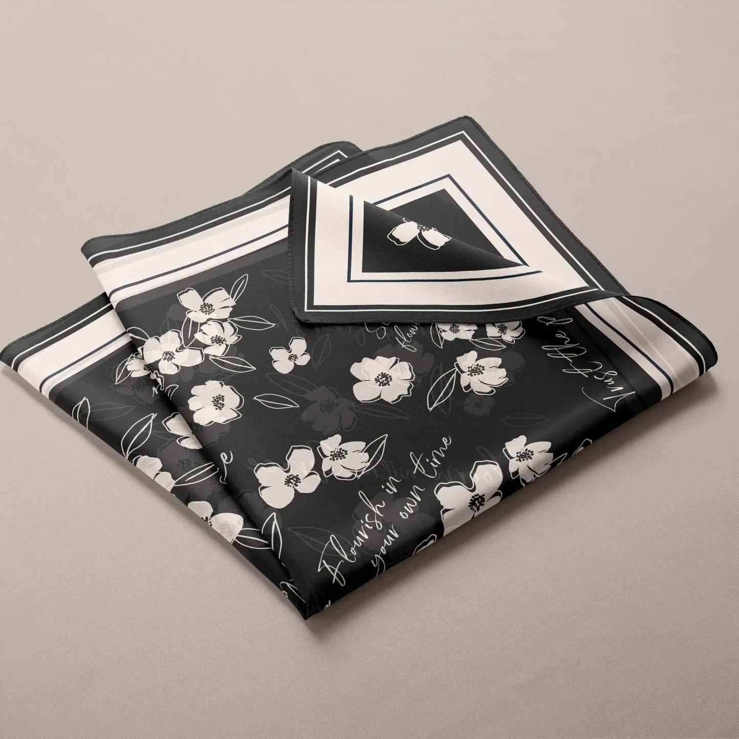 Folded black silk scarf featuring a white wildflower pattern and the inspirational phrase "Flourish in your own time"
