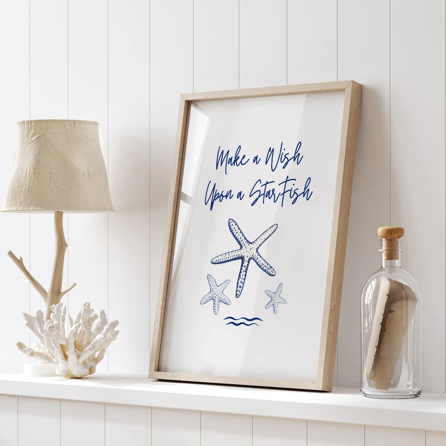 Coastal decor scene with framed starfish printable art, coral lamp, and glass bottle on a white shelf.