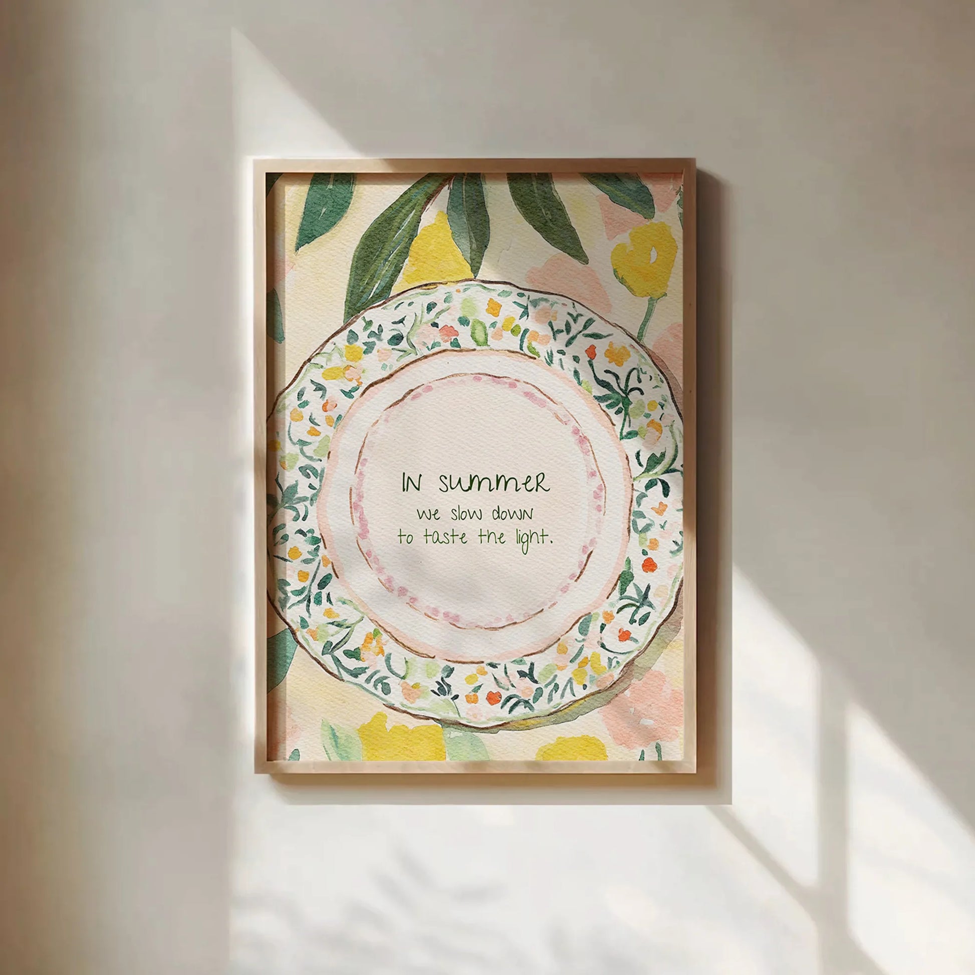Framed watercolor art of floral lemon plate with quote, styled in soft afternoon shadows on white wall.
