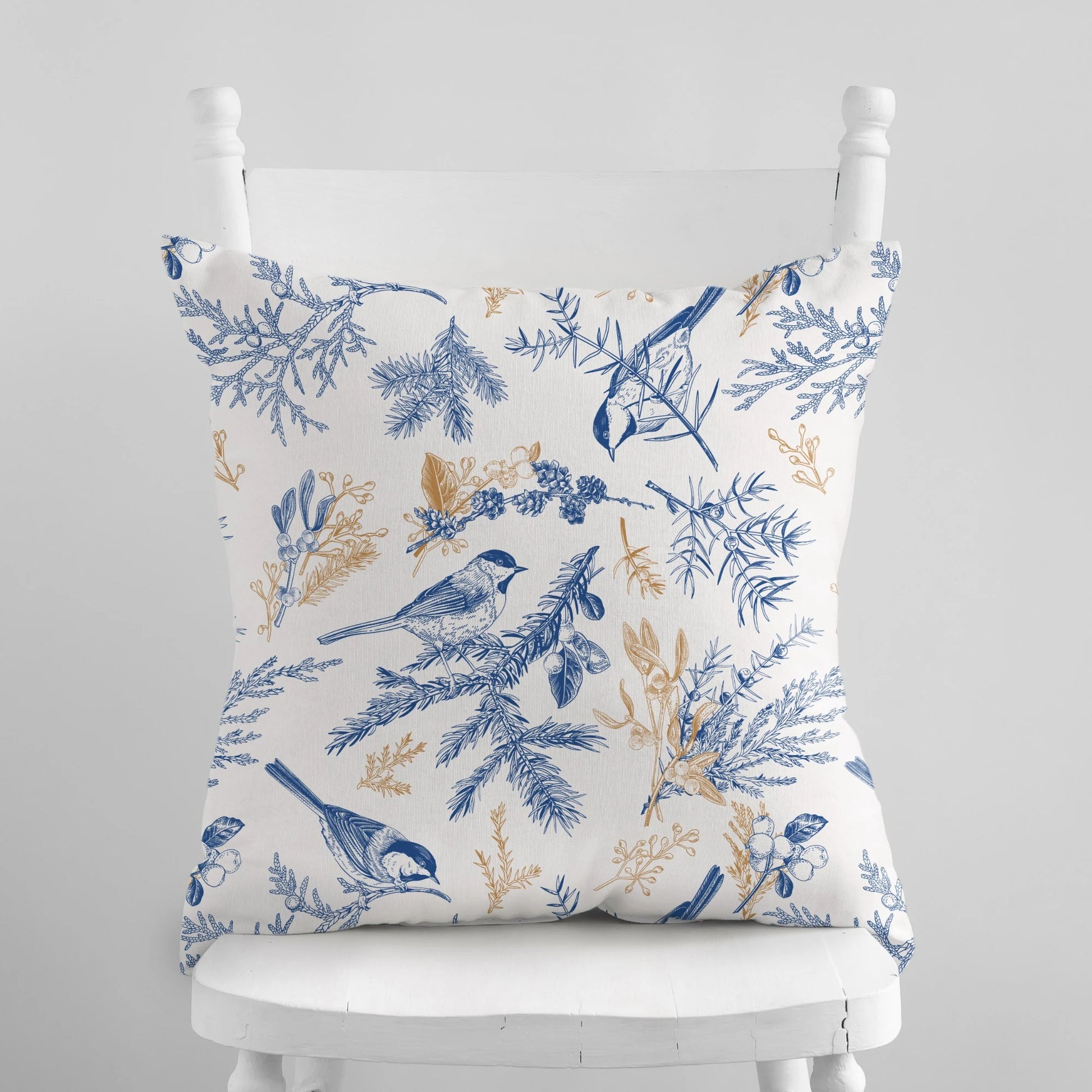 A square-shaped pillow with a vintage botanical blue and white pattern, featuring birds and winter berries, placed on a white chair.