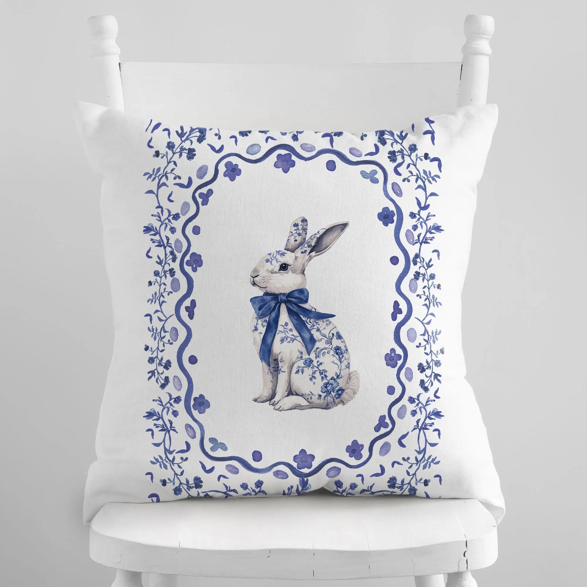 White pillow with a blue watercolor Chinoiserie rabbit wearing a bow, framed by a floral border.