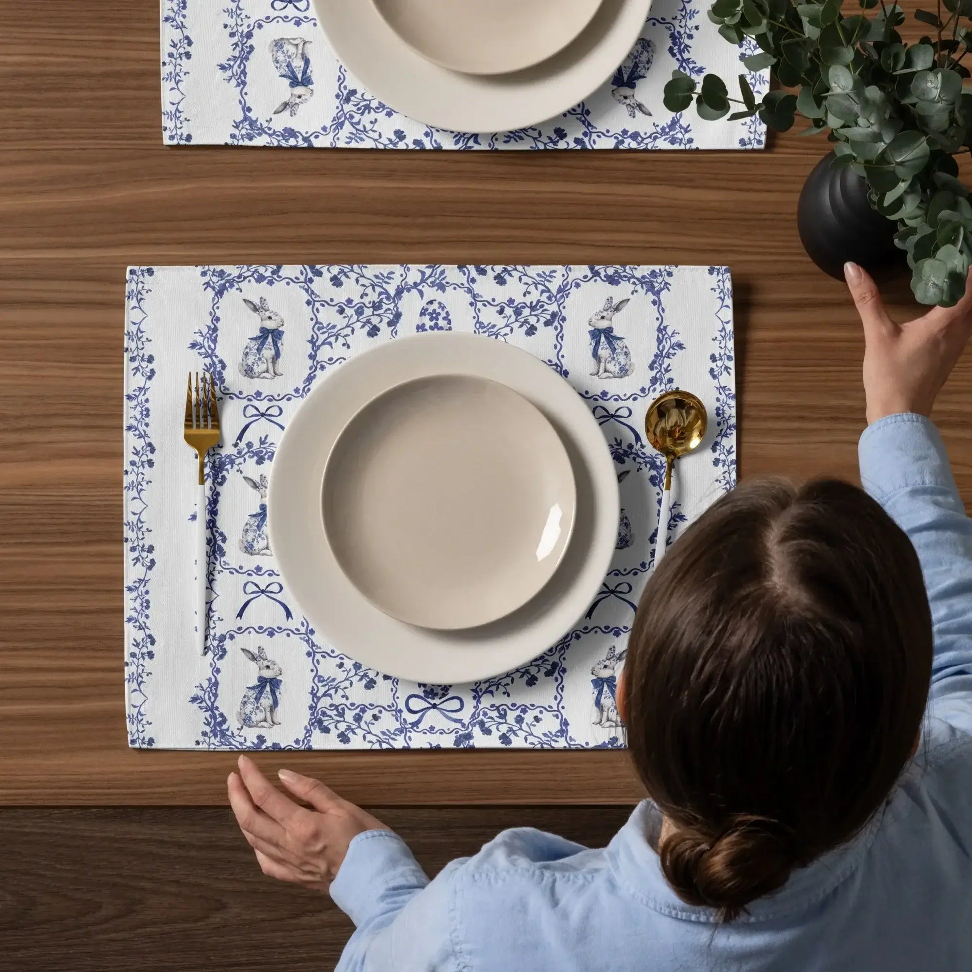 Overhead view of a blue and white placemat with a watercolor Chinoiserie rabbit and floral design on a wooden table being set with plates and gold flatware.