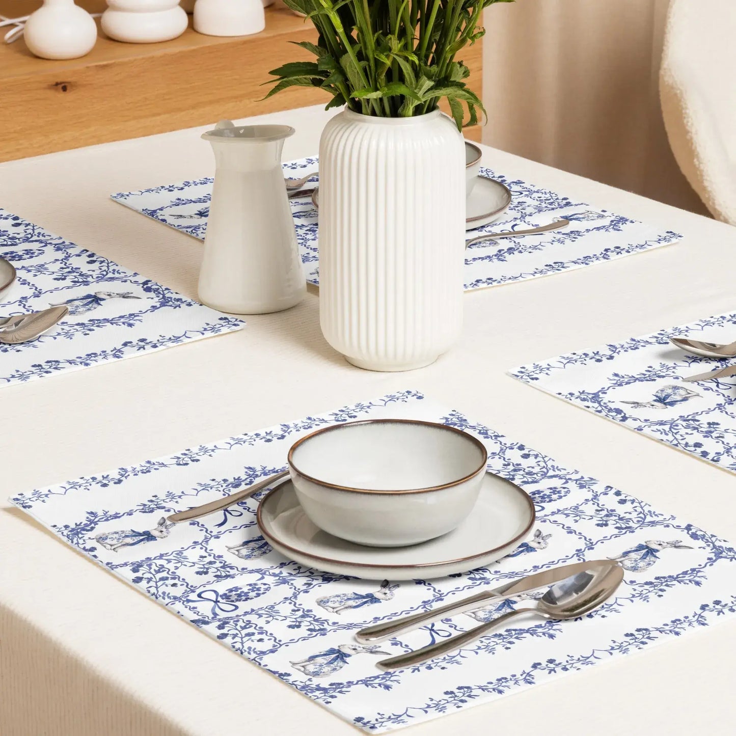Elegant blue and white placemats with a watercolor Chinoiserie rabbit and floral design on a white tablecloth with place settings and a vase of greenery.