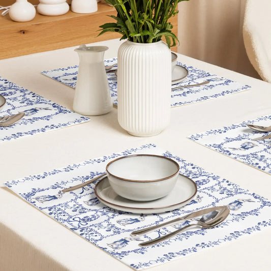 Elegant blue and white placemats with a watercolor Chinoiserie rabbit and floral design on a white tablecloth with place settings and a vase of greenery.