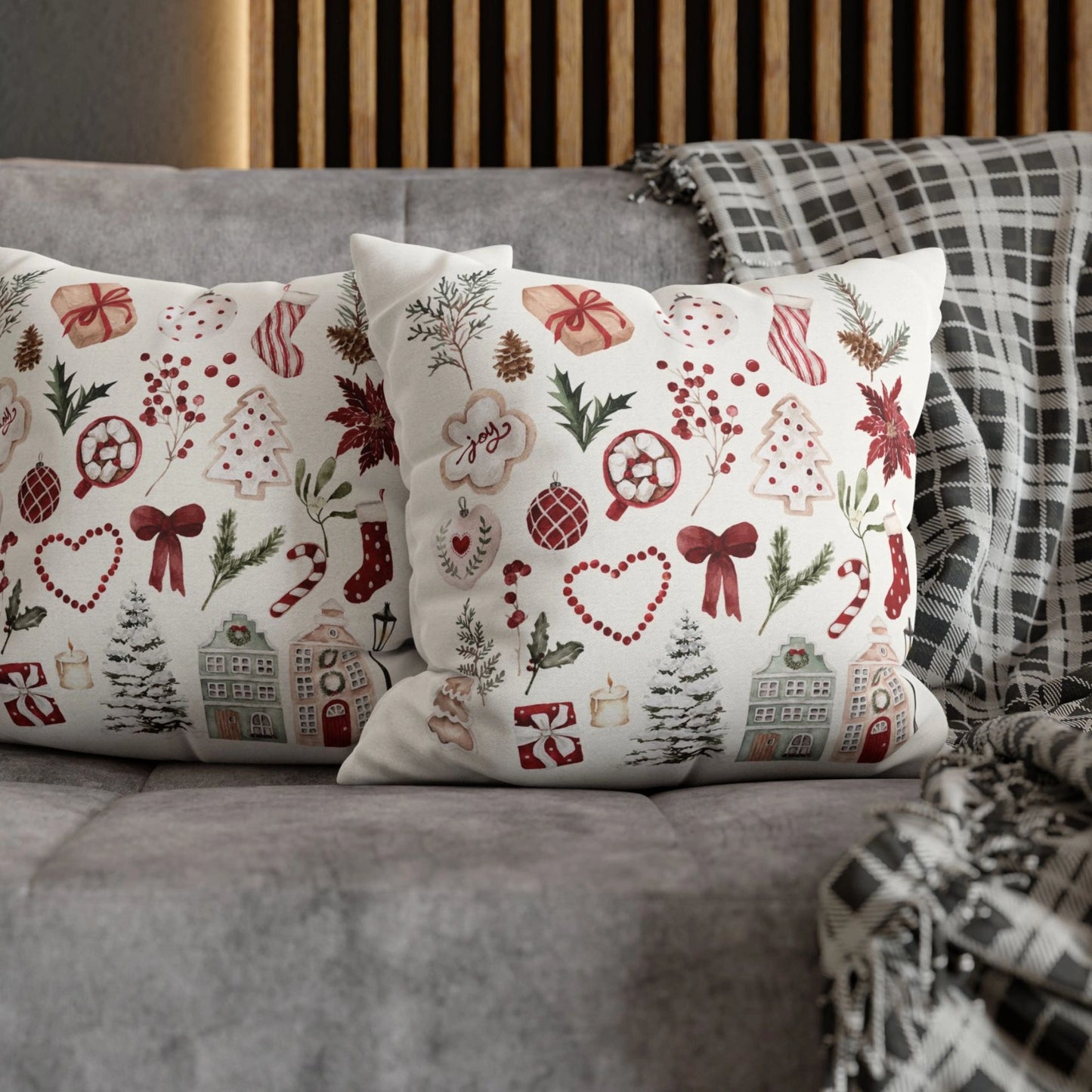 Pair of watercolor Christmas pillow covers on grey sofa – red and green festive design with poinsettias, pine, and holiday houses.