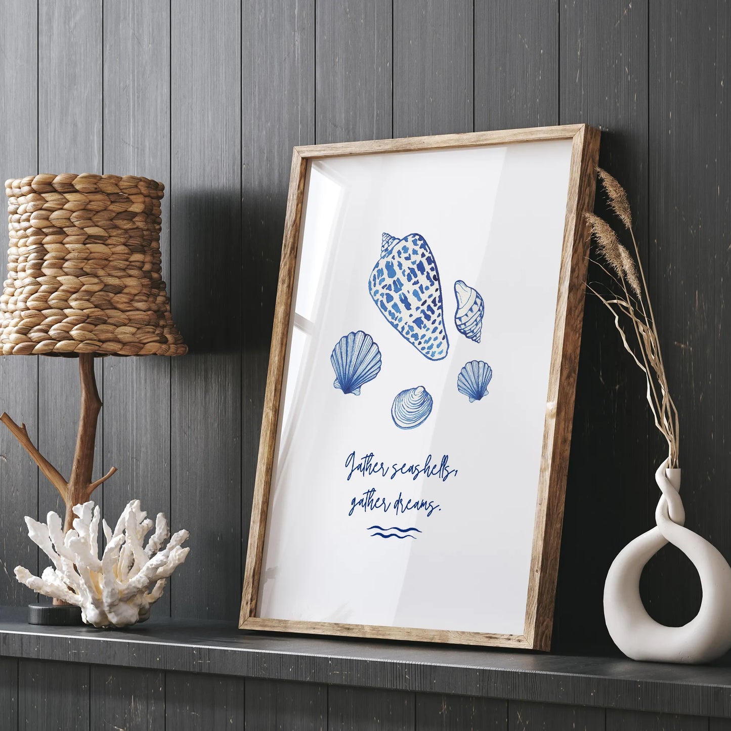 Watercolor seashell art print framed in rustic wood, styled on a dark coastal console table with woven lamp and coral sculpture decor.