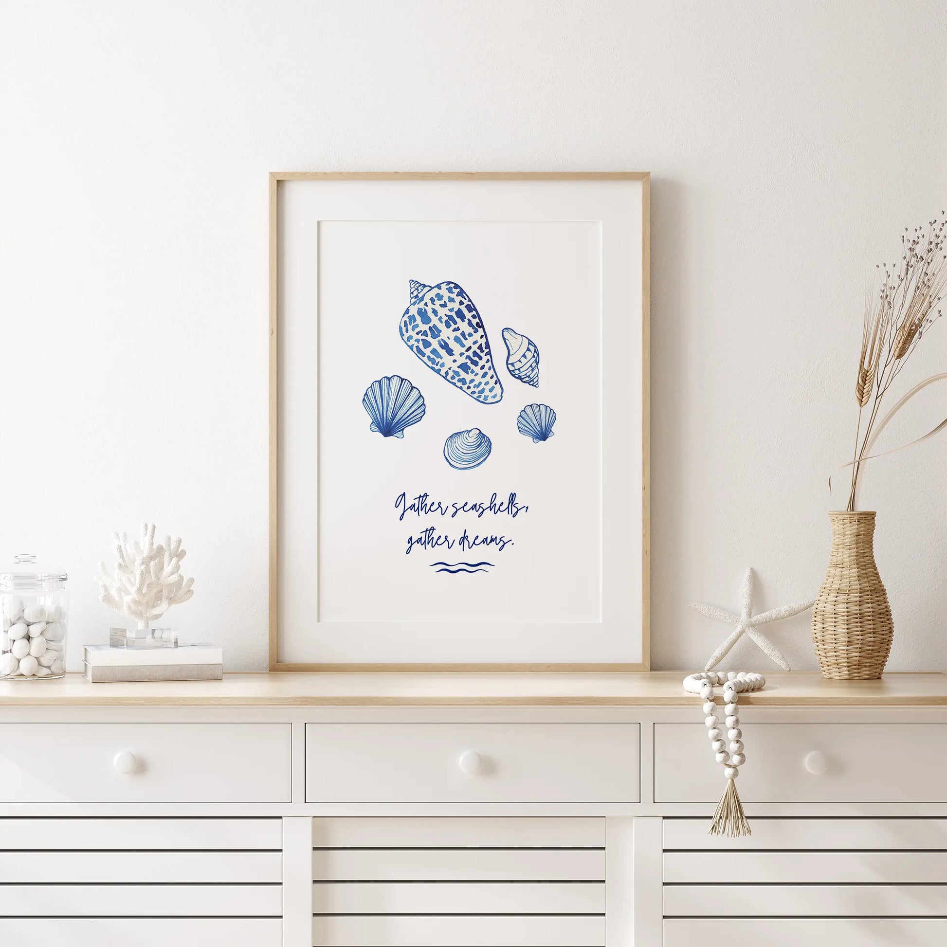 Coastal seashell wall art displayed in a white coastal-style room, featuring blue watercolor shells and "Gather seashells, gather dreams" inspirational quote.