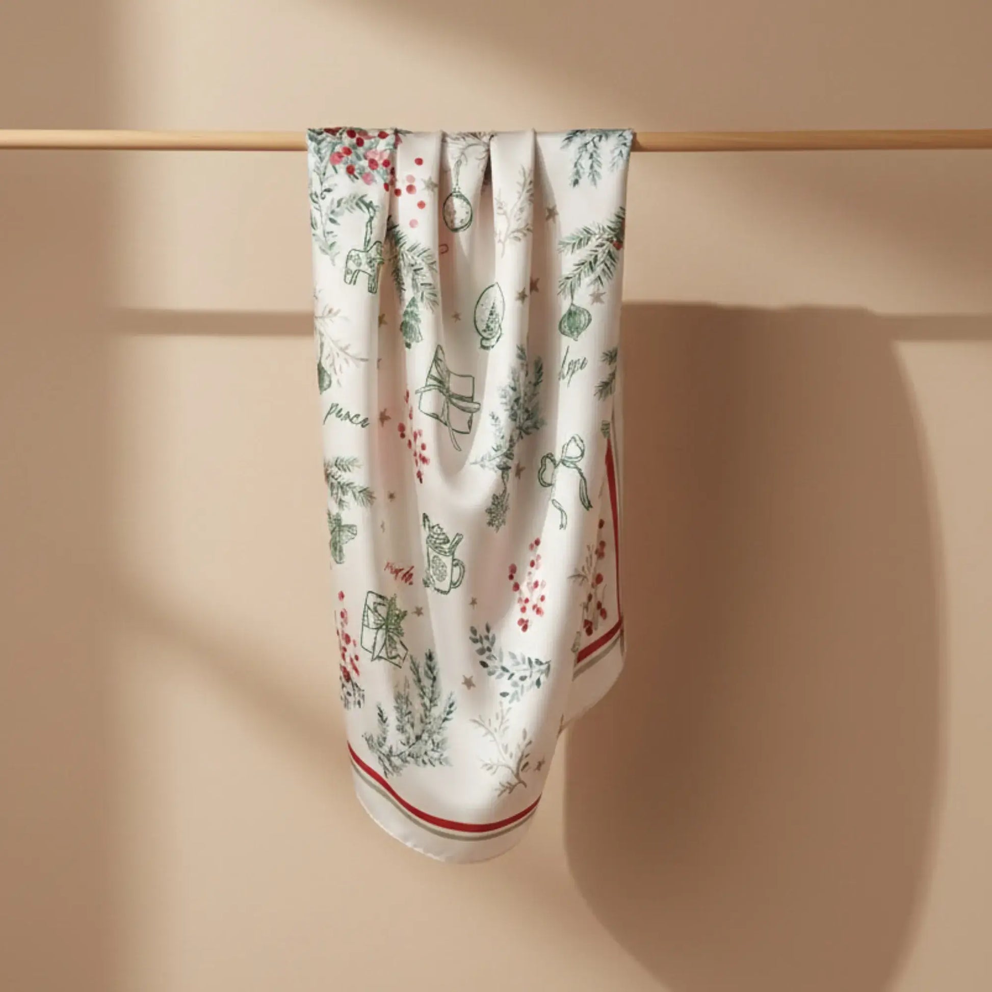 Winter Botanical Scarf hanging on a wooden rod, featuring hand-drawn winter designs and a festive red and green border.