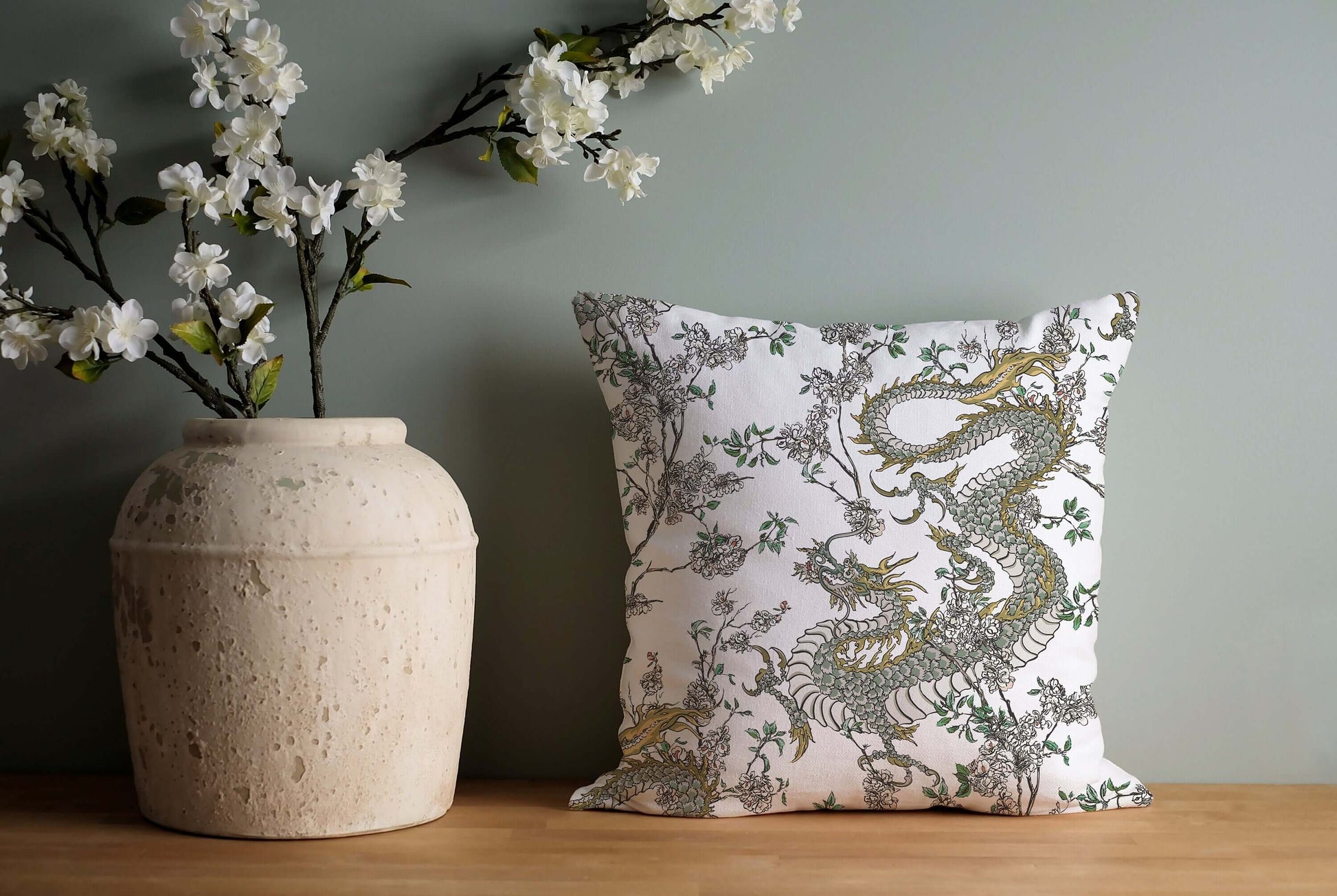 The pillow is placed on a wooden surface next to a light-colored vase holding white flowering branches. A light green wall is in the background.