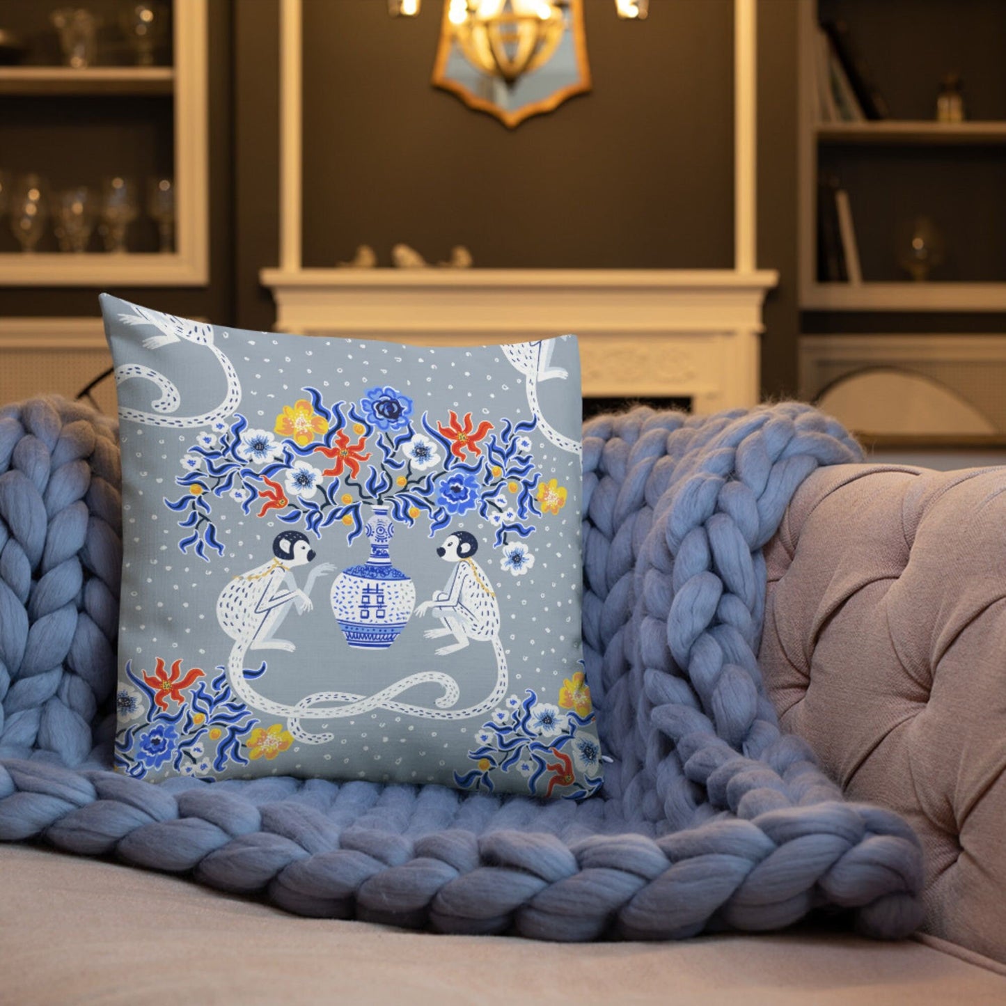Chinoiserie monkey throw pillow on grey sofa with knit blanket. Oriental design with blue pagoda vase and floral accents.
