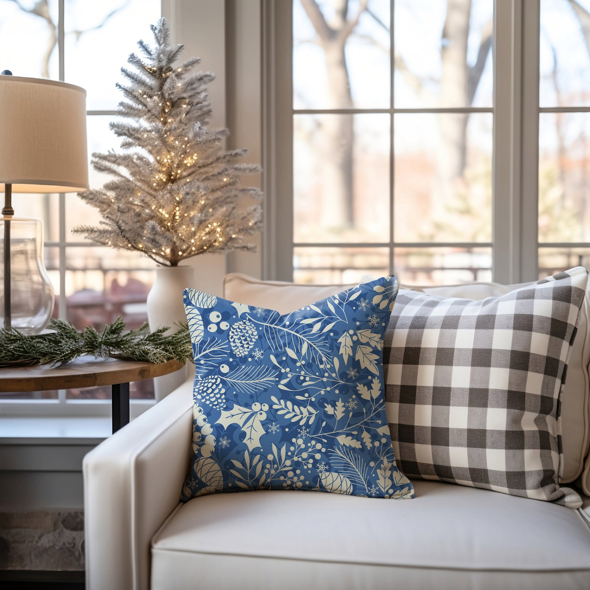 Light blue winter botanical pillow with cream pinecones and leaves on white sofa beside small Christmas tree